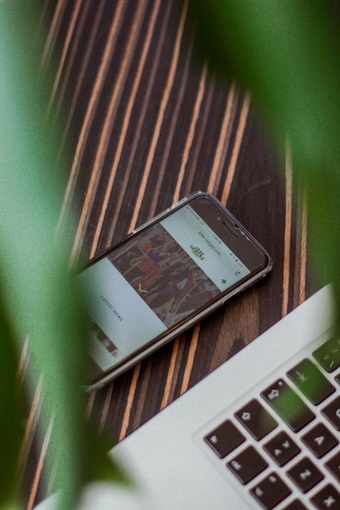 A smartphone displays a website on a wooden desk next to a laptop, surrounded by plants.